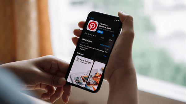 Photo shows a person holding a smartphone by a window. The phone screen shows the Pinterest App page on the App Store. This picture is part of the article on how to make money on Instagram.