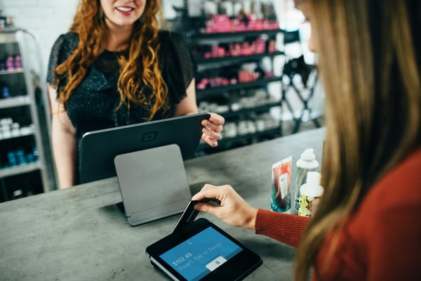 Photo shows a shop assistant behind a counter while a customer pays for a purchase by credit card, highlighting the importance of hiring additional help as a Black Friday marketing and sales strategy.