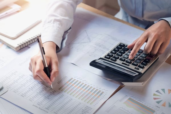 Photo shows a person's hands over a table. With the right hand, the person is holding a pen and writing on a sheet of paper, and with the other they're using the calculator. This photo helps demonstrate the importance of a break even analysis for small businesses.