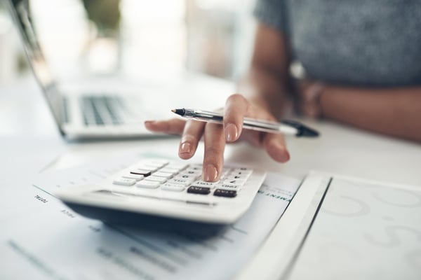 Photo shows a person sitting at a table, using a calculator and holding a pen. They're also working on their laptop. This photo alludes to the action of calculating break-even point.