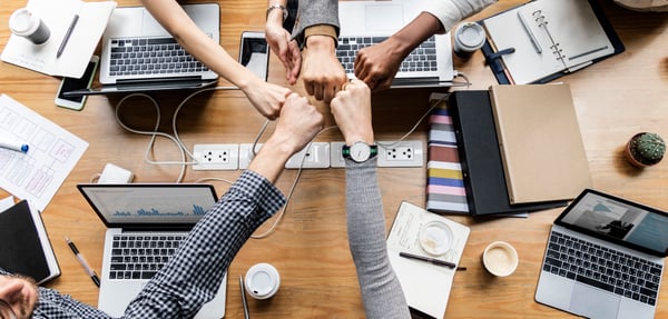 A desk filled with computers and office tools, with people touching fists representing team work.