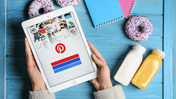 Picture shows a person's hands over a wooden table. The person is holding a tablet with the Pinterest app home screen open. There are donuts, notebooks and juice bottles scattered around the table. This image helps illustrate the blog post on how to make money on Pinterest.