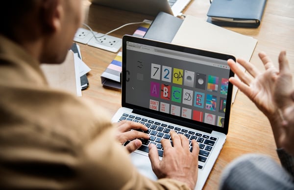 Photo that illustrates article on how to make enamel pins shows two people with their backs to the camera, facing a laptop. The camera focuses on the screen, which displays a series of images with different fonts and colors.