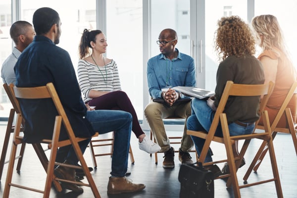 Photo shows six people sitting on folding chairs in a circle and talking. This photo represents focus groups, one of the methods used for market research.