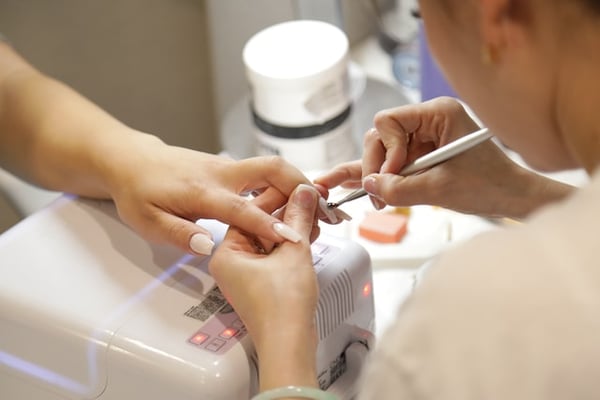 Photo shows a woman bending over a customer's hand while working on their gel nails. The customer's hand is placed on top of a nail dryer. This photo illustrates Shoplazza's article about the best dropshipping products.