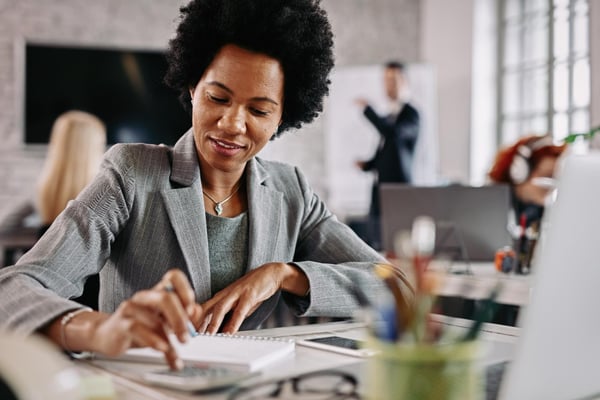 Photo shows a woman sitting at a table, wearing a suit, while using the calculator and writing down things on a notebook. The image alludes to calculating a business' break-even point.