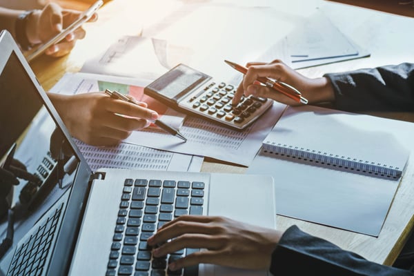 Photo focuses on a table and it shows two people sitting face to face. One of them is using a calculator and a laptop, while the other is looking at their phone and writing something down on a sheet of paper.  This photo helps illustrate how to do break even analysis.