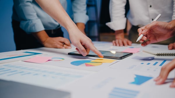 Picture focuses on a table, over which there are several pieces of paper and a tablet. There are a few people around the table, pointing at the graphics and charts on the paper. This picture was used to represent the market research required to sell the best dropshipping products.