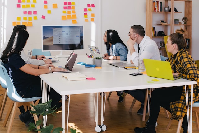 Photo shows five people sitting around a table and looking at a monitor screen. All of them have laptops in front of them, and there are post-it notes on the wall behind the monitor. This photo intents to illustrate doing a market research to start an online business in Canada.