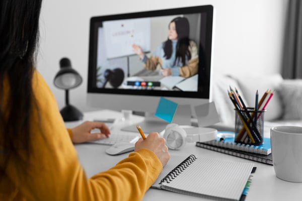 Photo from the digital products to sell online article shows a person sitting at a desk, taking notes on a notebook with a pencil. The person is staring at a monitor, and the screen shows a video call in progress. On the call, there is a person sitting down at a table, pointing at a white poster behind them.