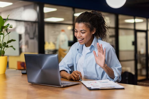 Photo shows a person sitting at a table in an office, waving at a laptop screen. This photo illustrates the virtual events section of the article about how to sell digital products online.