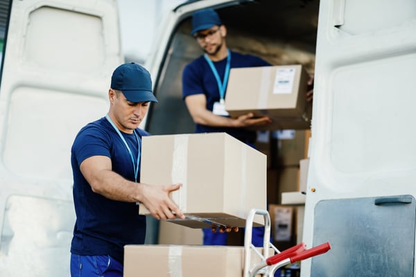 2 mens wearing a blue uniform removing boxes from a white car, representing outbound logistics.