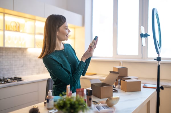 A woman in a kitchen with the table full of boxes, using a celphone and recording something with a ringlight to her youtube channel