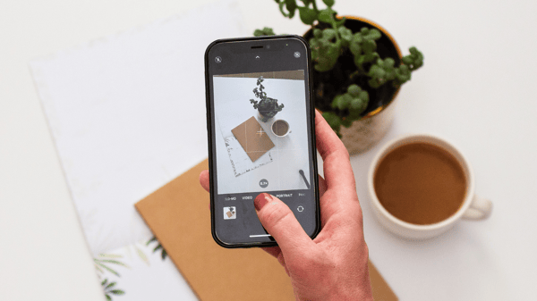 Photo shows a person holding a smartphone over a table, aiming the camera at the objects on the table. There is a potted plant, a coffee mug and some stationery on the table. This picture alludes to the importance of product photography when learning how to make money on Pinterest.