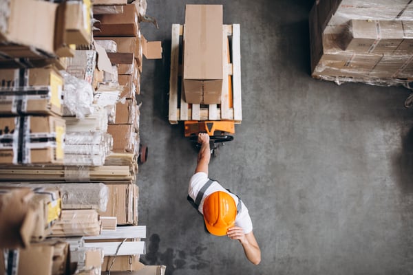 The top view of a warehouse, with a man using protection equipment while carrying boxes in a cargo cart, representing reverse logistics.