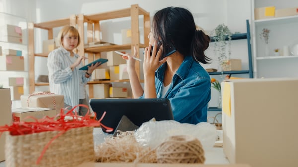 2 women in a store using tablets with boxes all around, to represent they're selling products online as a way to make money from home in Canada.