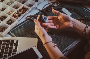 Photo shows a person holding a seashell-shaped pendant for a necklace. The person's hands are next to a laptop and a case full of beads. Photo illustrates article on how to start a jewelry business online.