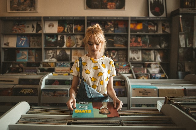 Photo illustrates blog post on starting an online business in Canada by selling second-hand products. In the photo, there is a person with long blond hair in a used record store, surrounded by shelves with records on display. The person is holding an Elvis Presley record.