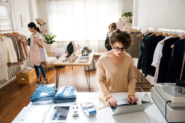 A showroom filled with clothes and the owner working on the laptop, representing how to decide on a business model for a clothing line.