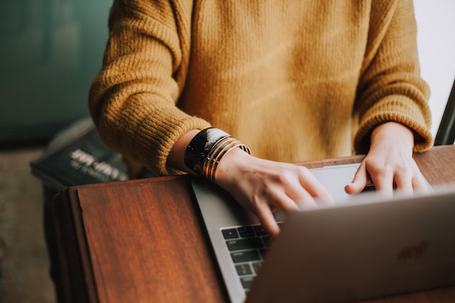 Photo illustrates article on starting an online business in Canada by showing someone working as a writer or translator. In the picture, there is a person sitting at a wooden table, typing on a laptop. The photo focuses on the person's arms and laptop. The person is wearing a knitted sweater and metal bracelets.