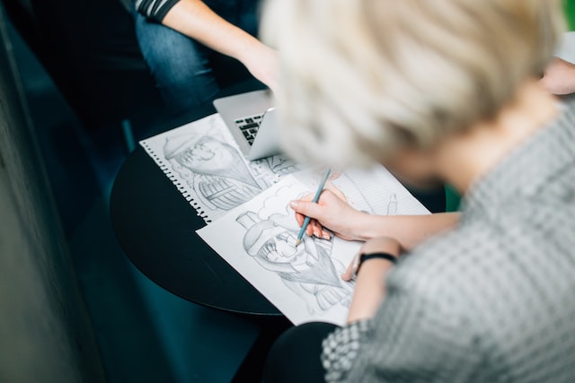 Picture that illustrates blog post to start an online business in Canada shows a person from the back, leaning over a table and drawing with pencil and paper. Opposite the drawer, there is another person using a laptop.