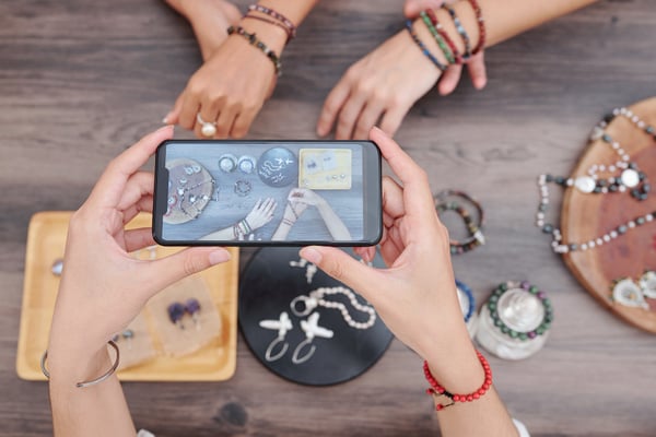 Picture shows a person using their smartphone to take a photo of several jewelry items to sell jewelry online. There are two people wearing bracelets and posing for the photo.