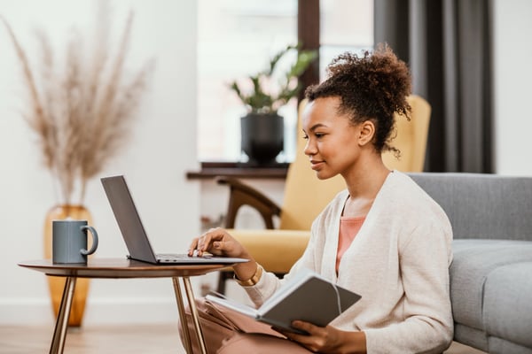 Photo shows a woman working from home, sitting on the floor and using a laptop. She's holding a notebook and she's looking at the screen. Photo illustrates how a person can start an online business in Canada as a virtual assistant.