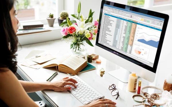 Photo shows a woman sitting at a desk, looking at a spreadsheet on a computer screen. This photo refers to the process of analyzing data for market research, as part of the article on what is market research.