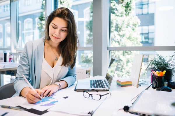 Photo from article entitled "What is opportunity cost?" shows a woman in an office setting, sitting at a desk, writing things down on a piece of paper with a laptop next to her.